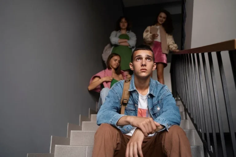 Teenage boy sitting on stairs looking distressed with a group of peers standing behind him, symbolizing youth mental health struggles.