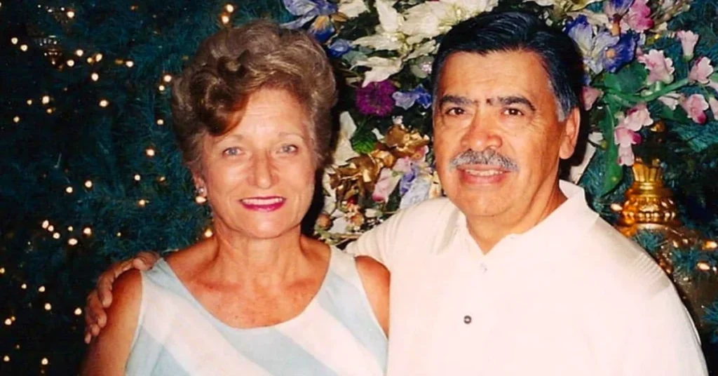 Camilla Consuelos and Saul Consuelos smiling together, the parents of actor Mark Consuelos, posing in front of decorative flowers and lights